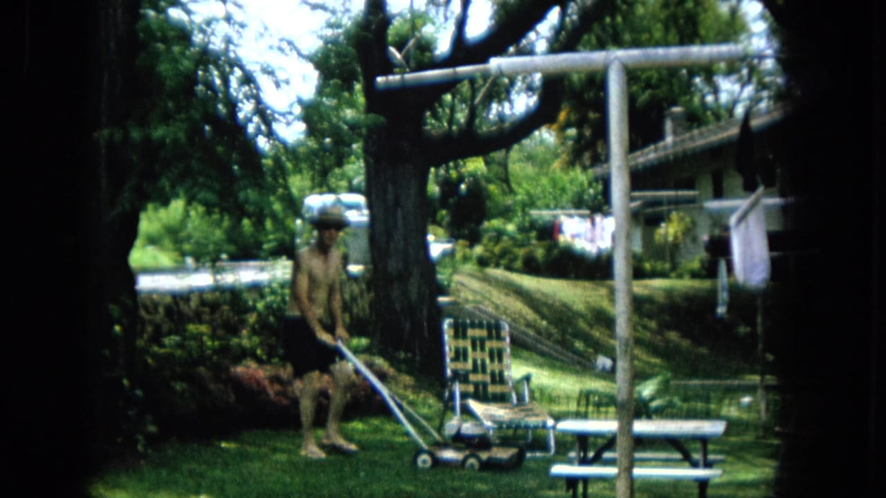 1965 U.S. backyard footage frame with a man using a push mower in dappled sunlight near a woven lawn chair, picnic table, laundry line, shrubs, and tree trunk.