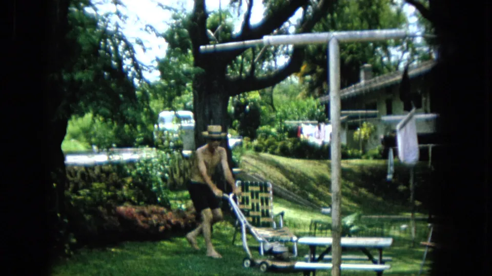 1965 U.S. home-movie frame of a shirtless man guiding a push mower past a woven chair and picnic table, with shrubs, laundry, and a large tree in the yard.