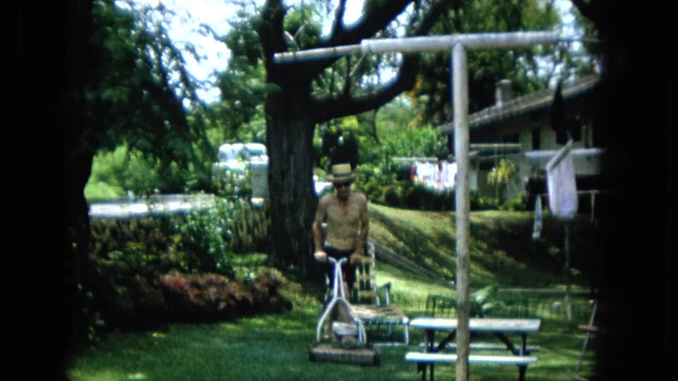 1965 U.S. backyard frame showing a shirtless man in a straw hat facing the camera behind a push mower, with a picnic table, lawn chair, clothesline, shrubs, and shade tree visible.