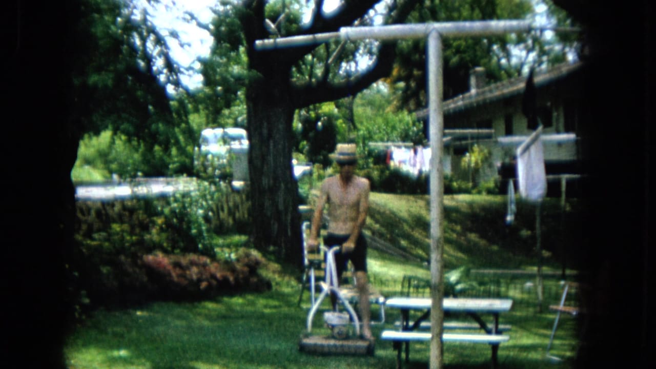1965 United States Super 8 frame showing a shirtless adult man in a straw hat and dark shorts mowing a green suburban backyard lawn with a picnic table and clothesline nearby.