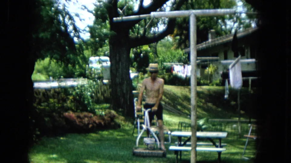 1965 United States Super 8 frame showing a shirtless adult man in a straw hat and dark shorts mowing a green suburban backyard lawn with a picnic table and clothesline nearby.