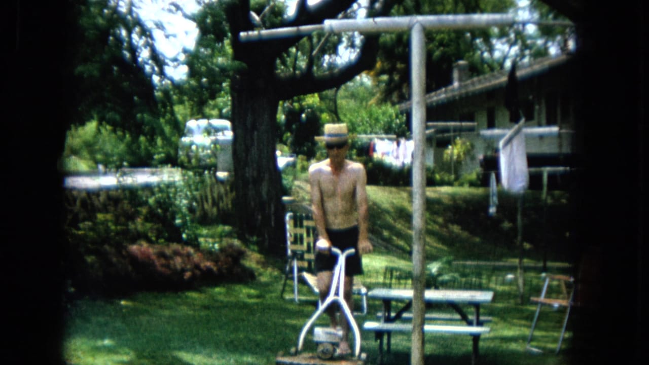 Man with a straw hat stands behind a manual mower near a picnic table in a backyard.