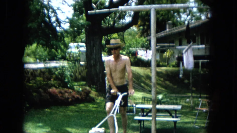 1965 United States Super 8 still showing the man closer to the camera as he pushes a white-handled reel mower through a sunny backyard lawn.