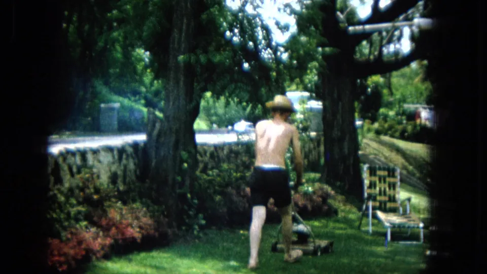 1965 United States backyard frame showing the man from behind as he pushes the manual mower away under trees, with patio furniture and a house visible in the background.