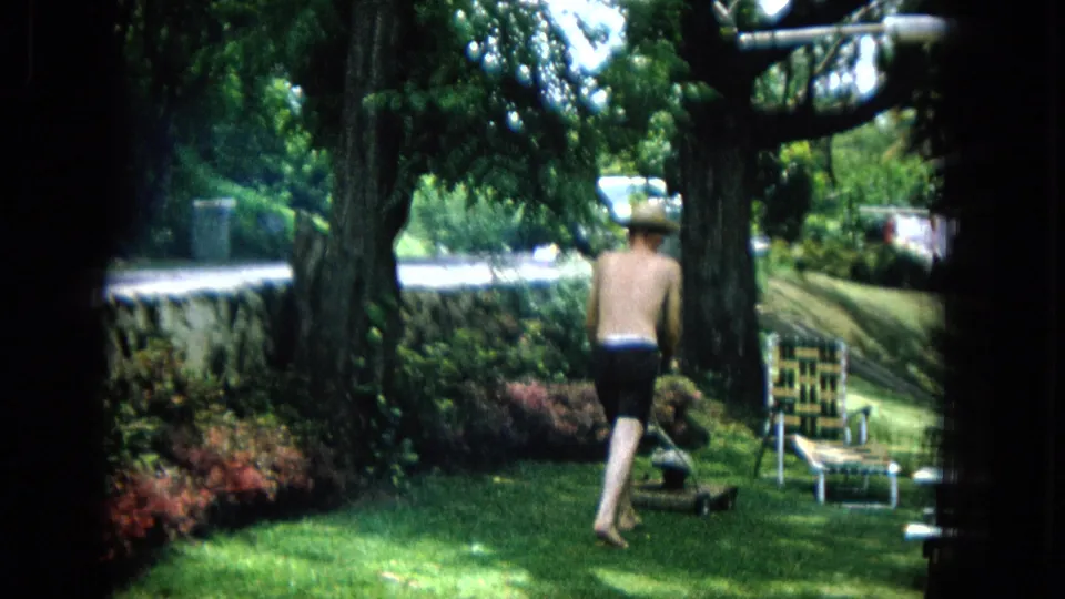 In a 1965 United States backyard scene, a shirtless man in dark shorts and straw hat walks toward a manual mower near a woven lawn chair under shade trees.
