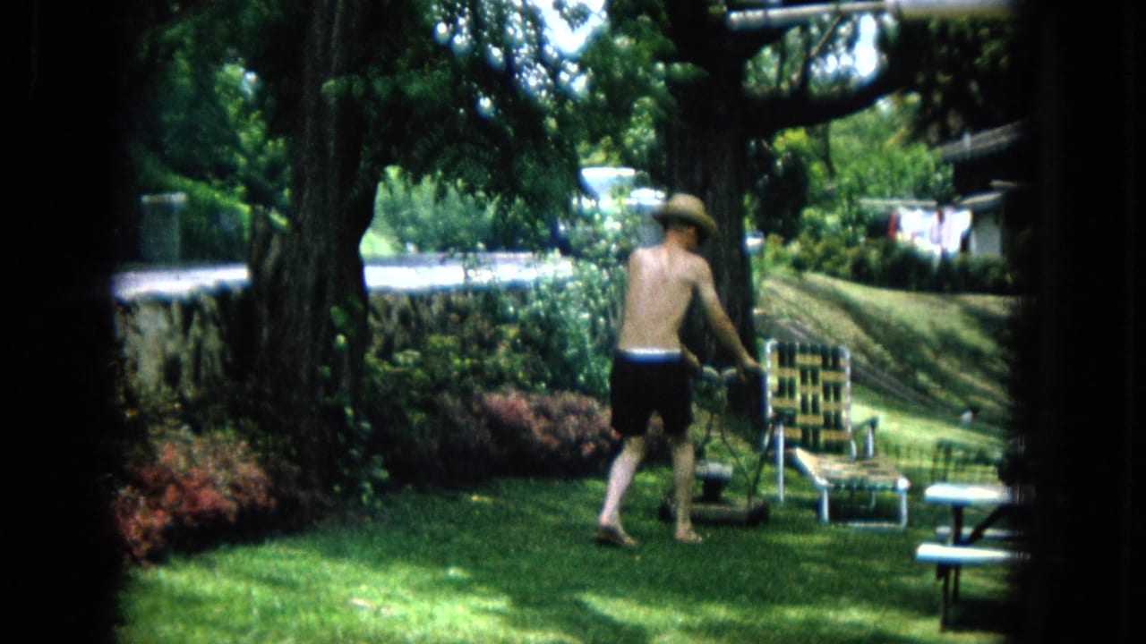 The man pauses beside the manual mower on the lawn, with a woven chair, shrubs, low wall, and dappled tree shade visible in the suburban yard.