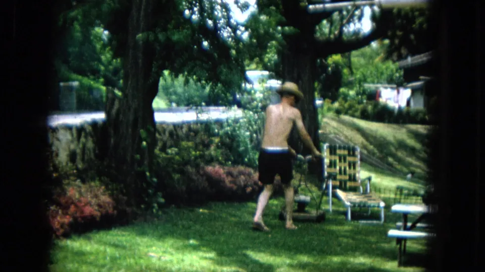The man pauses beside the manual mower on the lawn, with a woven chair, shrubs, low wall, and dappled tree shade visible in the suburban yard.