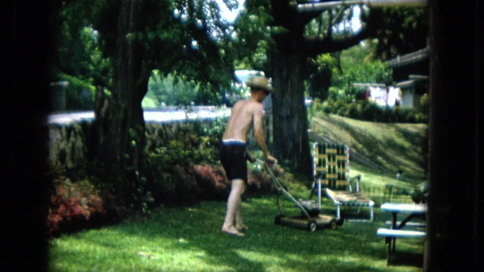 The shirtless man guides the reel mower through the grass, framed by mature trees, shrubs, a low wall, and a woven lawn chair in the background.