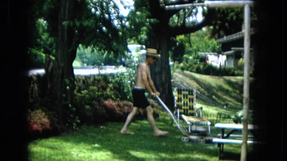 The man moves the push mower across the backyard lawn, passing a woven chair and picnic table beneath tree shade in the 1965 home-movie footage.