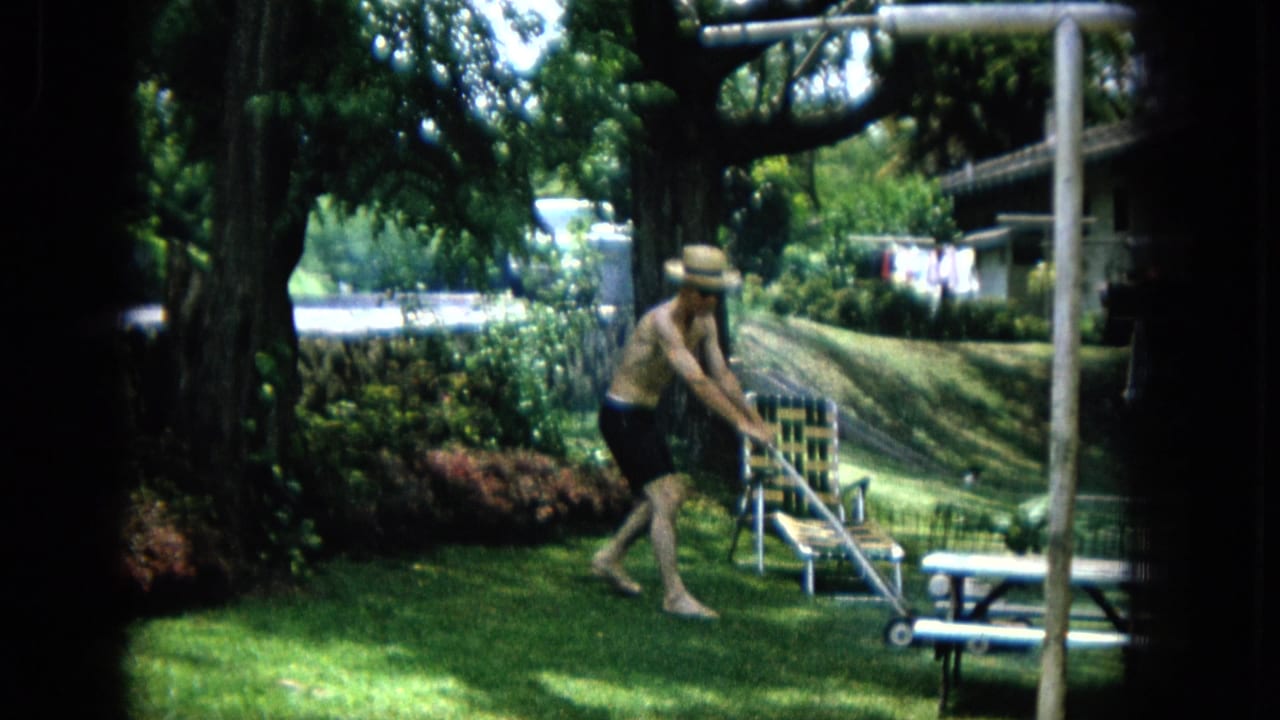 1965 Super 8 frame of a shirtless adult man in a straw hat mowing a green backyard lawn in the United States.