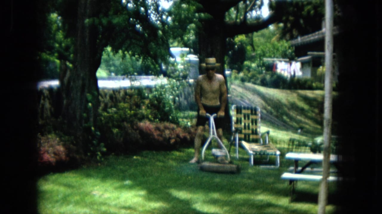 Man stands behind a push mower near lawn chairs under trees.