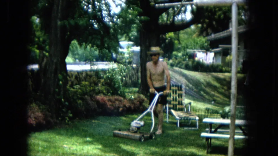 1960s home-movie frame showing a shirtless man moving a push mower across a shaded green lawn.