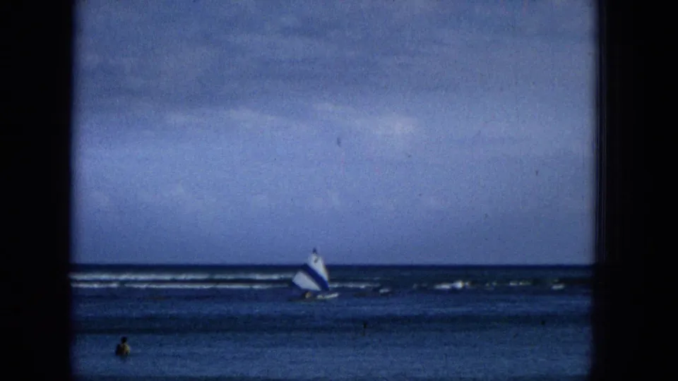 Distant small sailboat on ocean water, with a swimmer or figure visible closer to the camera in a blue-toned Super 8 frame.