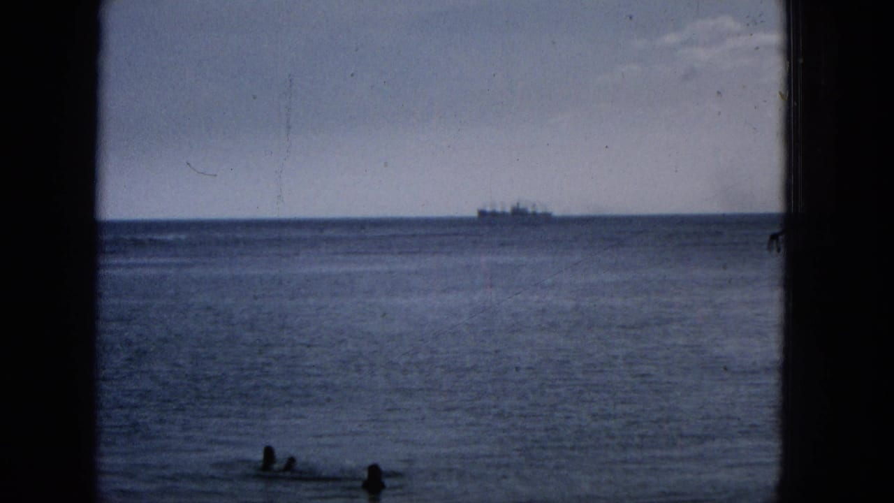 1965 Hawaii home-movie frame showing an ocean horizon through dark window or structural framing, with two swimmers near shore and a ship far offshore.