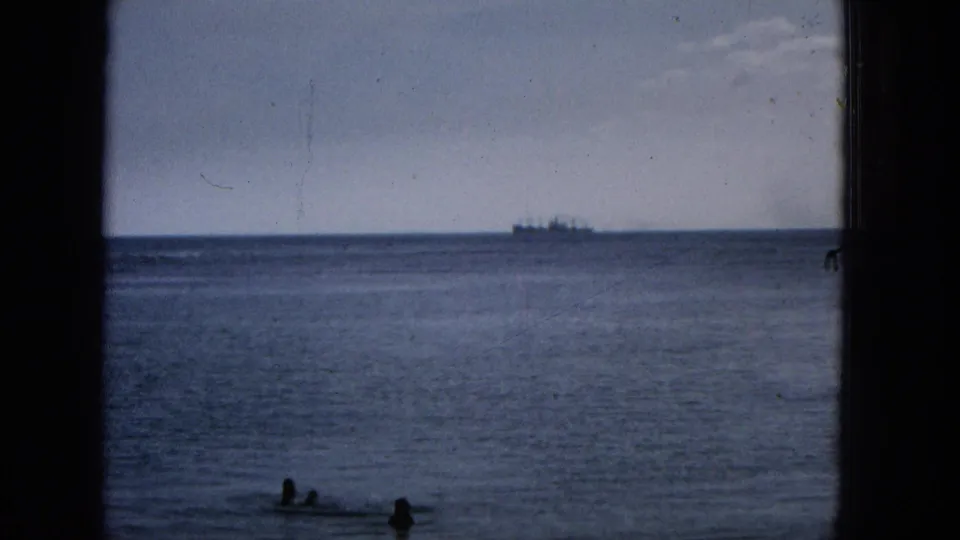 1965 Hawaii home-movie frame showing an ocean horizon through dark window or structural framing, with two swimmers near shore and a ship far offshore.