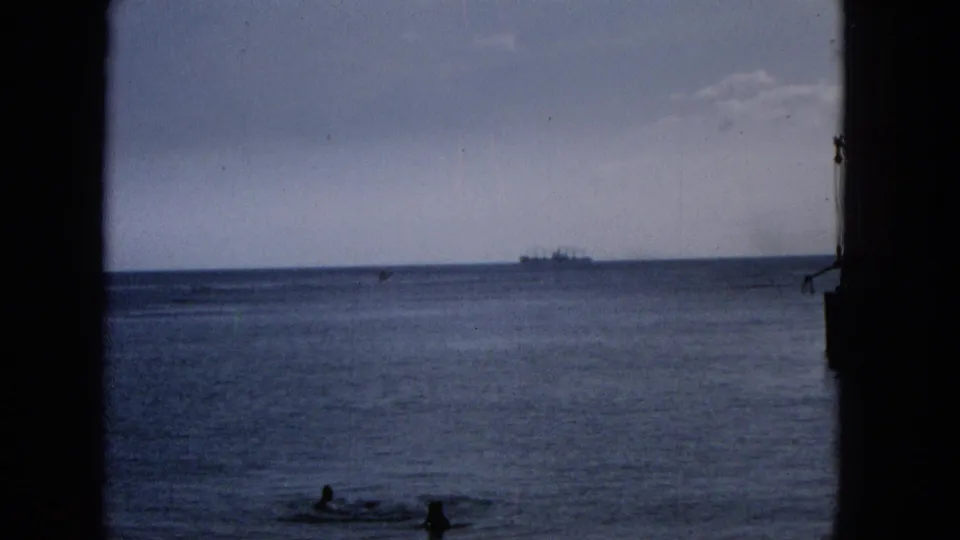 1965 Hawaii shoreline view through dark framing, with swimmers in the foreground water and a ship far out on the ocean horizon.