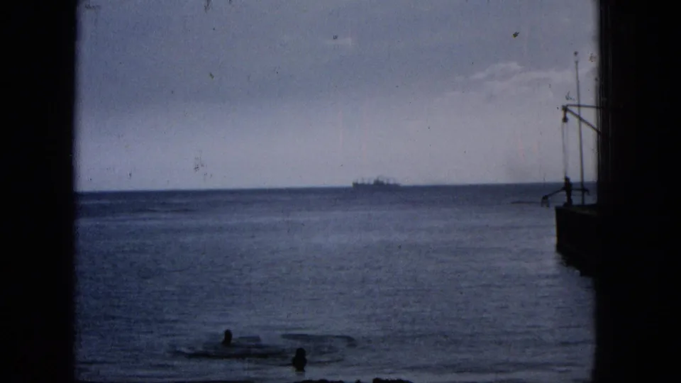 1965 Hawaii coastal home-movie frame with swimmers in nearshore water, a distant ship on the horizon, and dark window or dock-like framing at the edges.