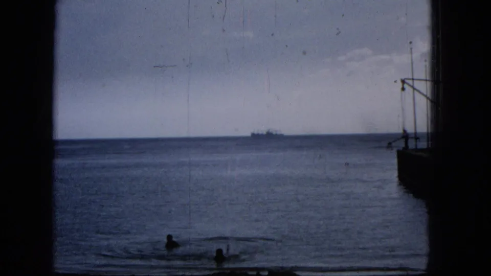 1965 Hawaii home-movie view of swimmers near shore, an offshore ship on the horizon, and dark structural framing around the seascape.