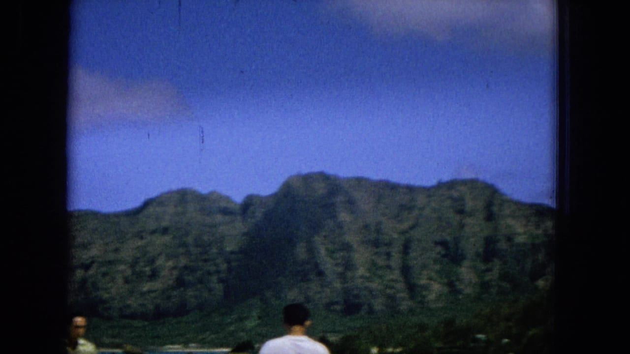1965 8mm Hawaii home-movie frame showing a green mountain ridge above a shoreline, with two casually dressed men partially visible in the foreground.