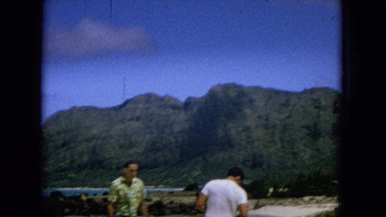 Two men stand near a beach with mountains and water behind them.