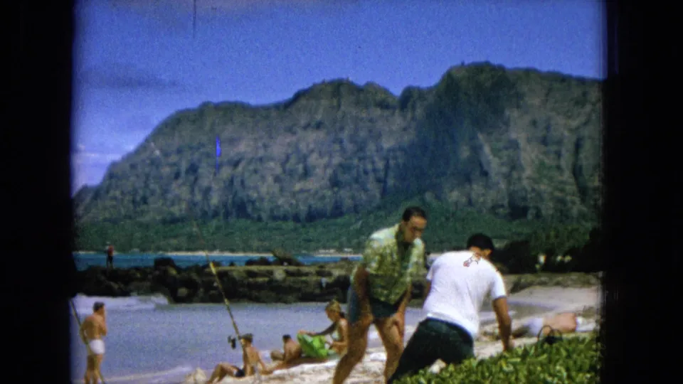 1965 8mm Hawaii shoreline scene showing beachgoers on the sand, one person fishing, and two men walking or standing near the foreground with mountains behind them.
