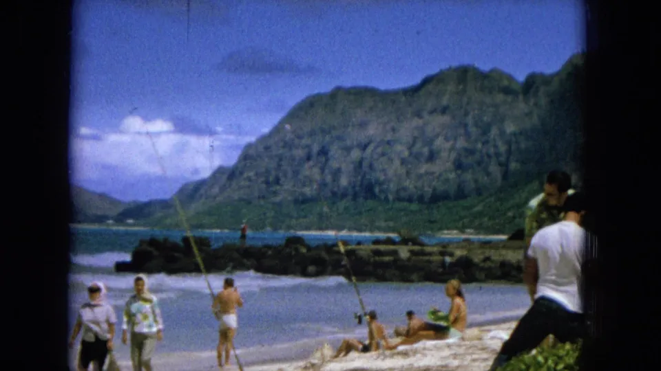 1965 8mm Hawaii beach frame with scattered people sunbathing, fishing, and walking along a sandy shoreline, with rocky water and green mountains in the distance.