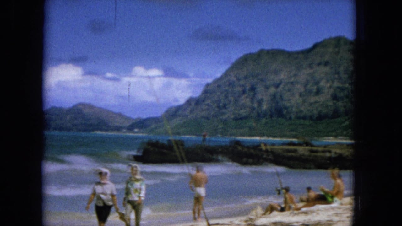 1965 Hawaiian beach scene with several people on the sand, waves rolling in, rocky shoreline, and a steep green mountain backdrop.