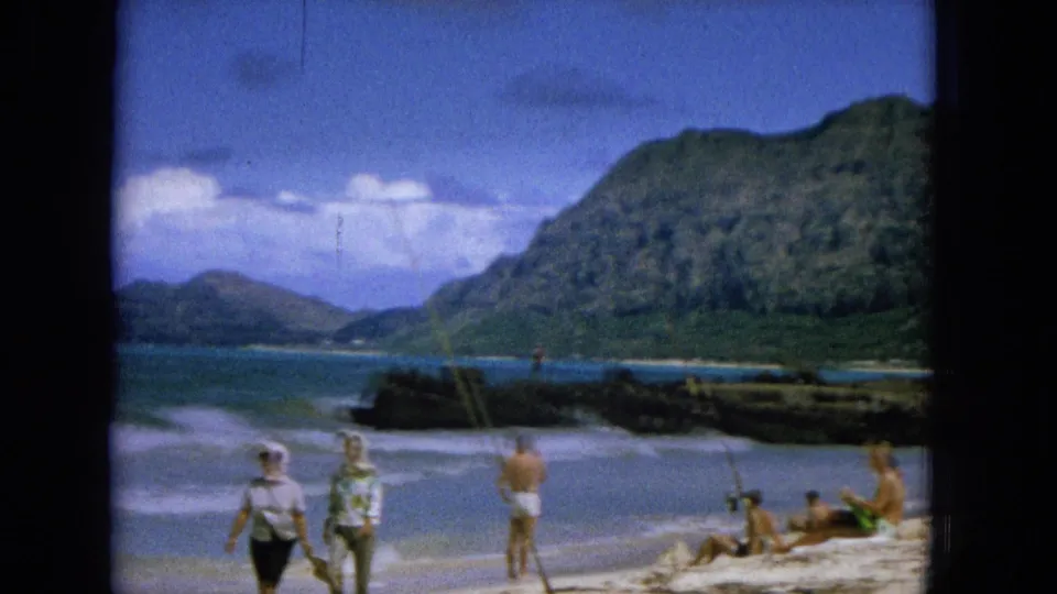 1965 Hawaiian beach scene with several people on the sand, waves rolling in, rocky shoreline, and a steep green mountain backdrop.