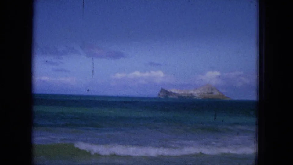 1965 Hawaiian ocean cutaway showing low surf in the foreground, open water, and a rocky offshore landform beneath scattered clouds.