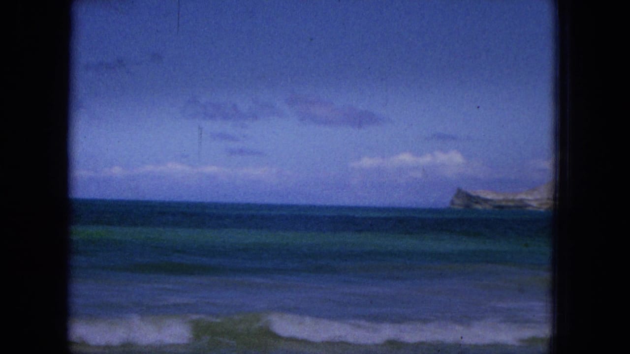1965 Super 8 home-movie frame from Hawaii showing a cloudy ocean horizon, low waves, dark film borders, and a distant headland or island shape near the right edge.
