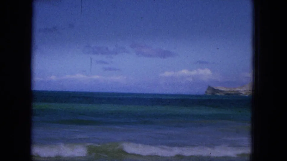 1965 Super 8 home-movie frame from Hawaii showing a cloudy ocean horizon, low waves, dark film borders, and a distant headland or island shape near the right edge.