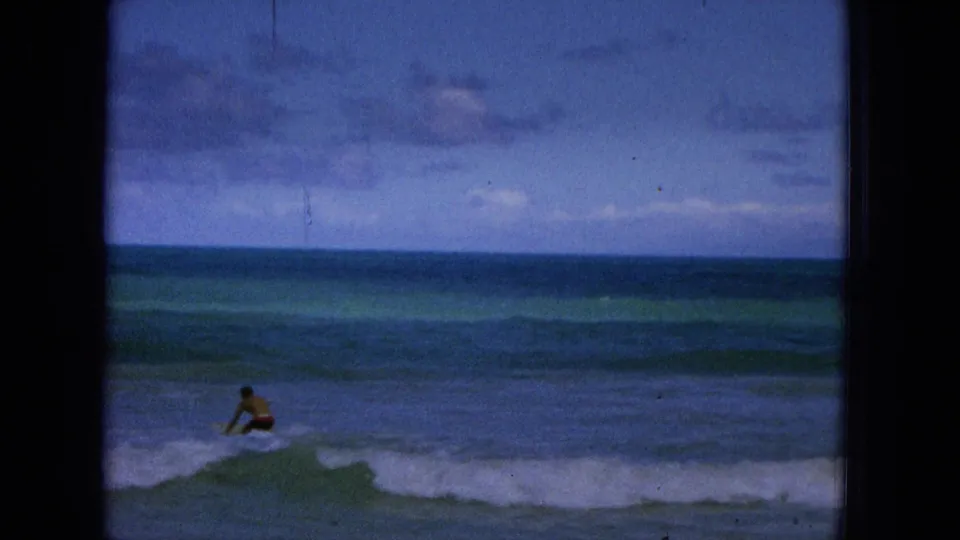 1965 Super 8 beach frame from Hawaii with a distant surfer or swimmer moving through shore break beneath a cloudy blue sky and open ocean horizon.