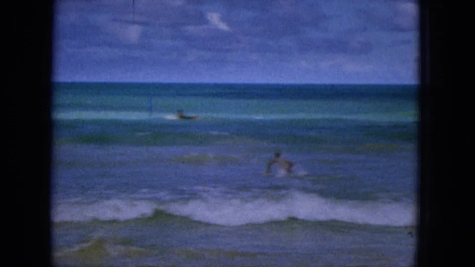 1965 Super 8 frame from Hawaii showing distant beachgoers or surfers in the water as small waves break across a soft, blue-green ocean view.