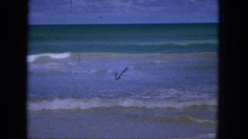 1965 Super 8 home-movie frame from Hawaii showing low surf washing toward a sandy beach, with a small indistinct object or figure near the waterline.