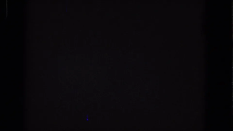 Dark 1965 Super 8 frame showing grain, a small blue speck, and faint border artifacts rather than a readable scene.