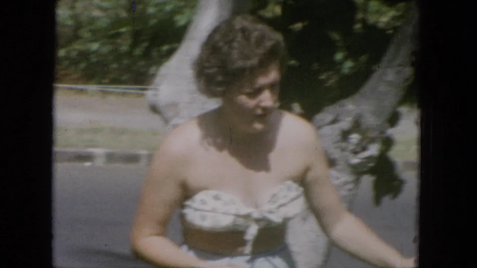 Adult woman in casual 1960s-style clothing walks beside a large pale tree trunk in daylight, with grass and a paved path behind her.