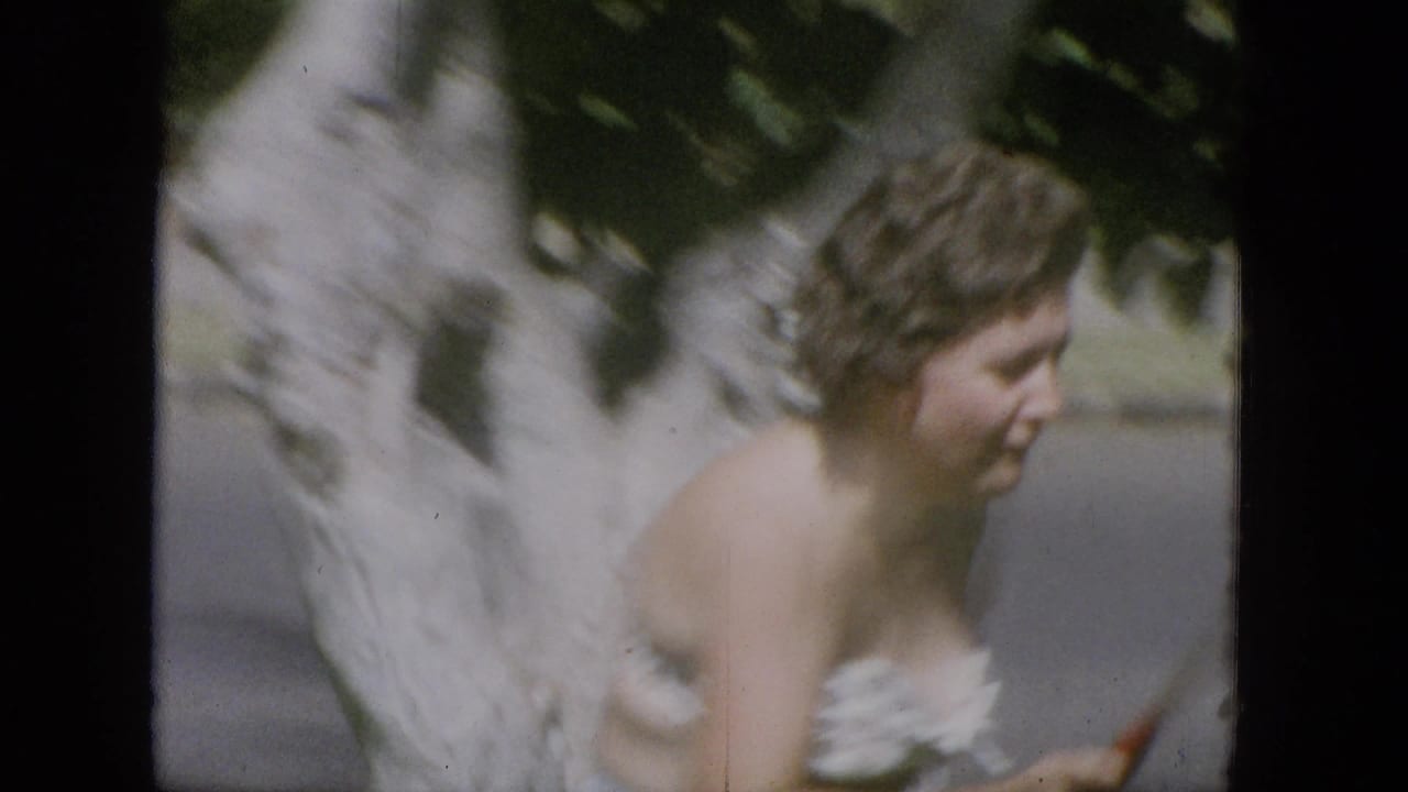 Woman passes a large tree trunk while holding a tennis racket at the edge of the frame.