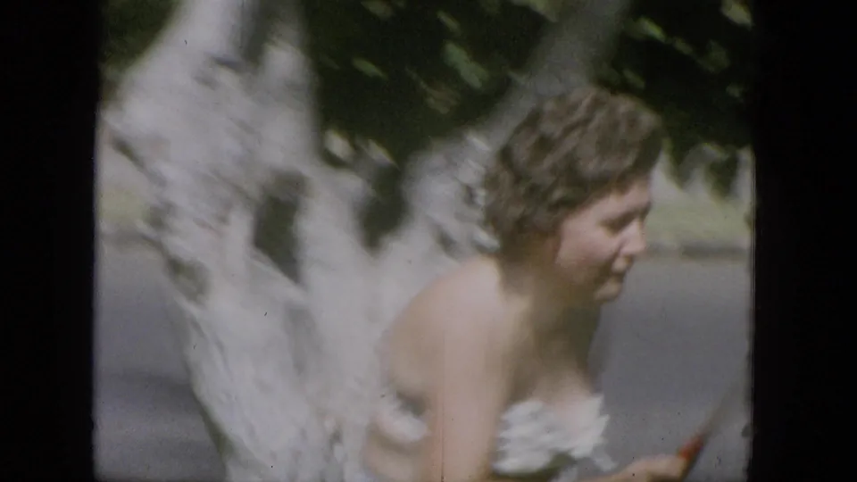 Adult woman moves past a large pale tree trunk in a sunlit outdoor area, with part of a tennis racket visible near the right edge.