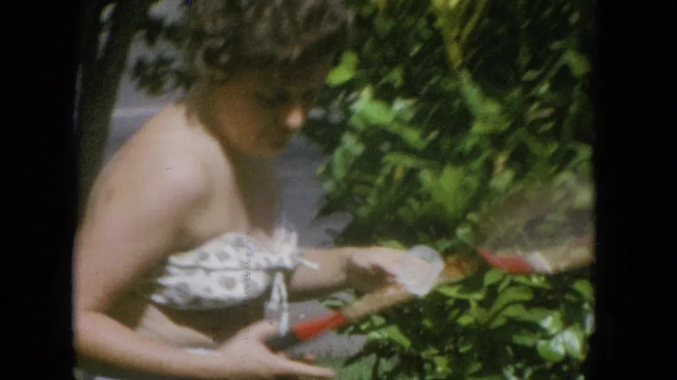 Adult woman in a strapless patterned top holds a tennis racket near leafy green shrubs, with the soft color and blur of 8mm home-movie footage.