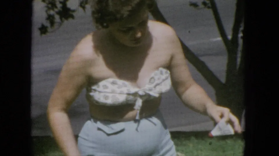 Adult woman walks through a grassy outdoor area beside a tree, carrying a tennis racket with a paved path visible in the background.