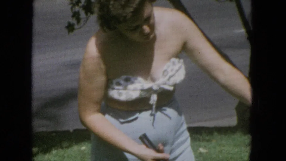 A woman in a patterned strapless top and light pants stands on a United States lawn in 1965 home-movie footage, holding a badminton racket near trees and a road.