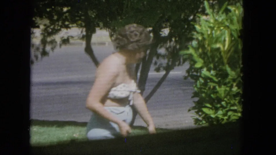 A side view shows the woman in a yard with a badminton racket, shrubs, trees, and a paved road visible in the background of the 1965 home-movie frame.