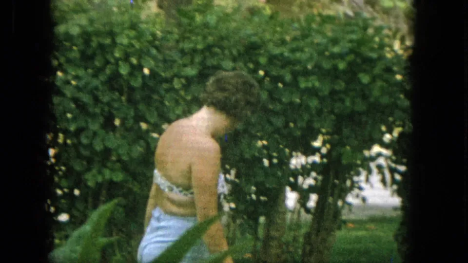 The woman bends forward on the lawn near dense shrubs and trees, with her badminton activity continuing in soft, grainy 1965 United States 8mm footage.