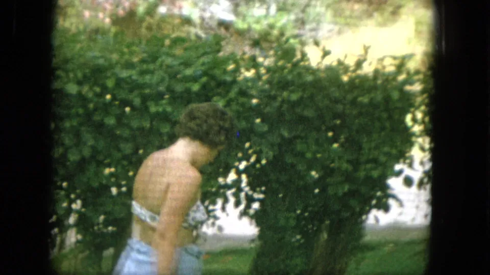 A shirtless man stands or moves near tropical-looking greenery in archival 1965 home-movie footage associated with a Hawaii family vacation.