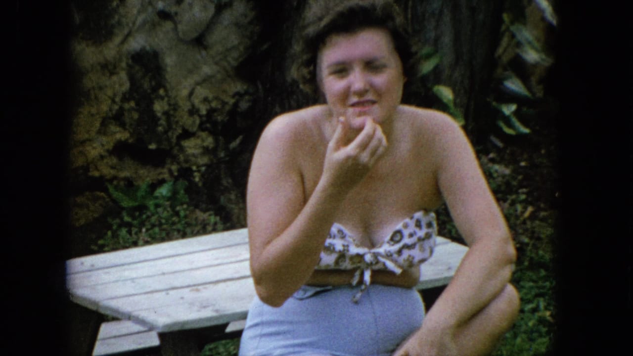 A woman in casual 1960s summer clothing sits by a wooden picnic table in a lush outdoor setting, smiling toward the camera.