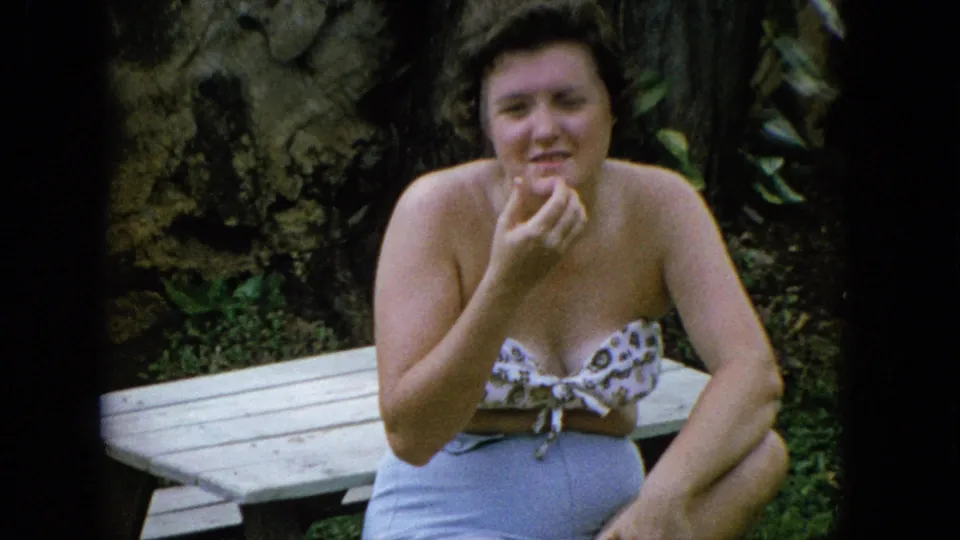 A woman in casual 1960s summer clothing sits by a wooden picnic table in a lush outdoor setting, smiling toward the camera.