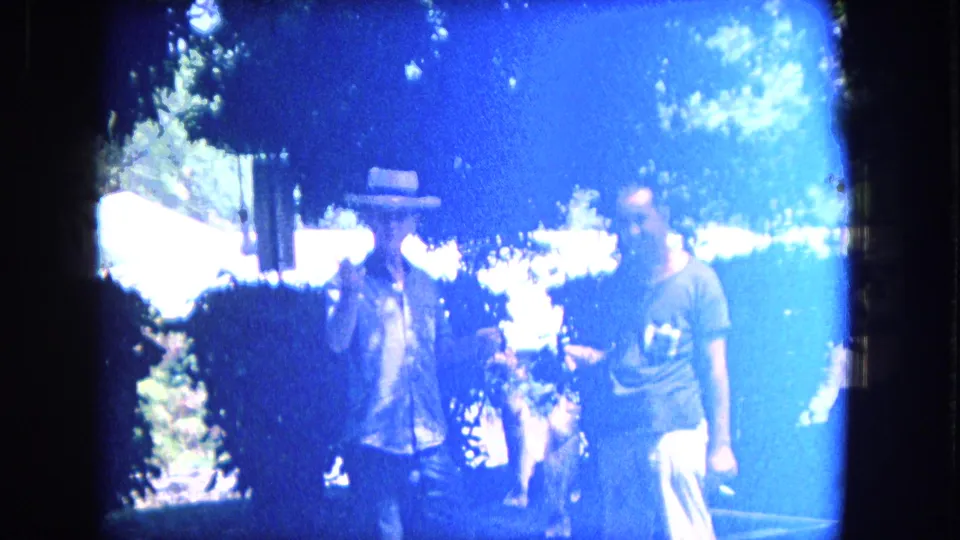 Two men appear under trees in a heavily flared outdoor home-movie frame, with bright overexposure and foliage surrounding them.