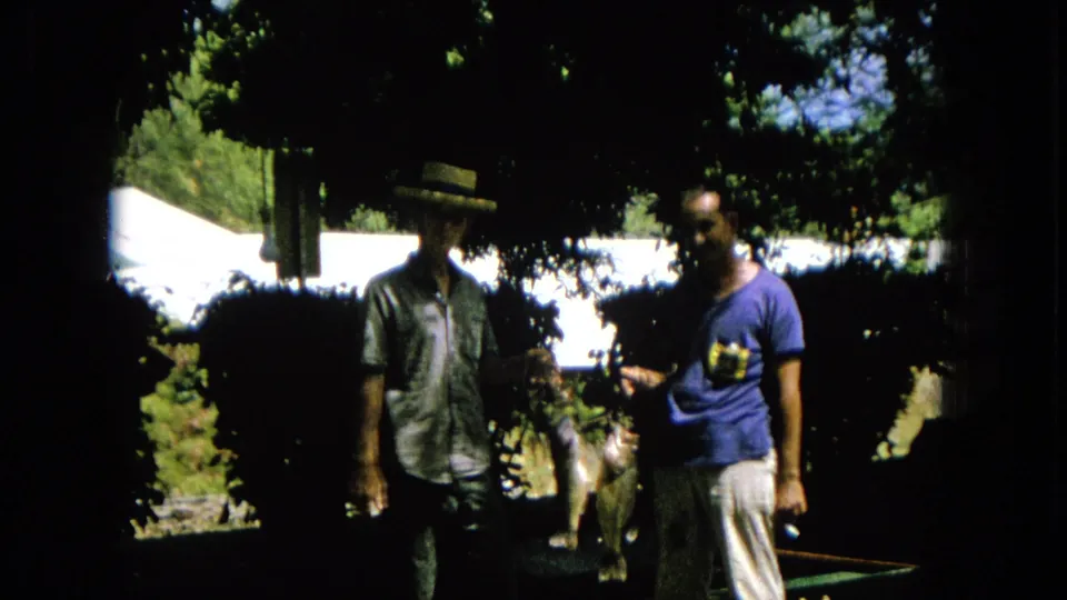 Two casually dressed men stand in deep shade beneath trees, with a white tent or canopy visible behind them in the outdoor scene.