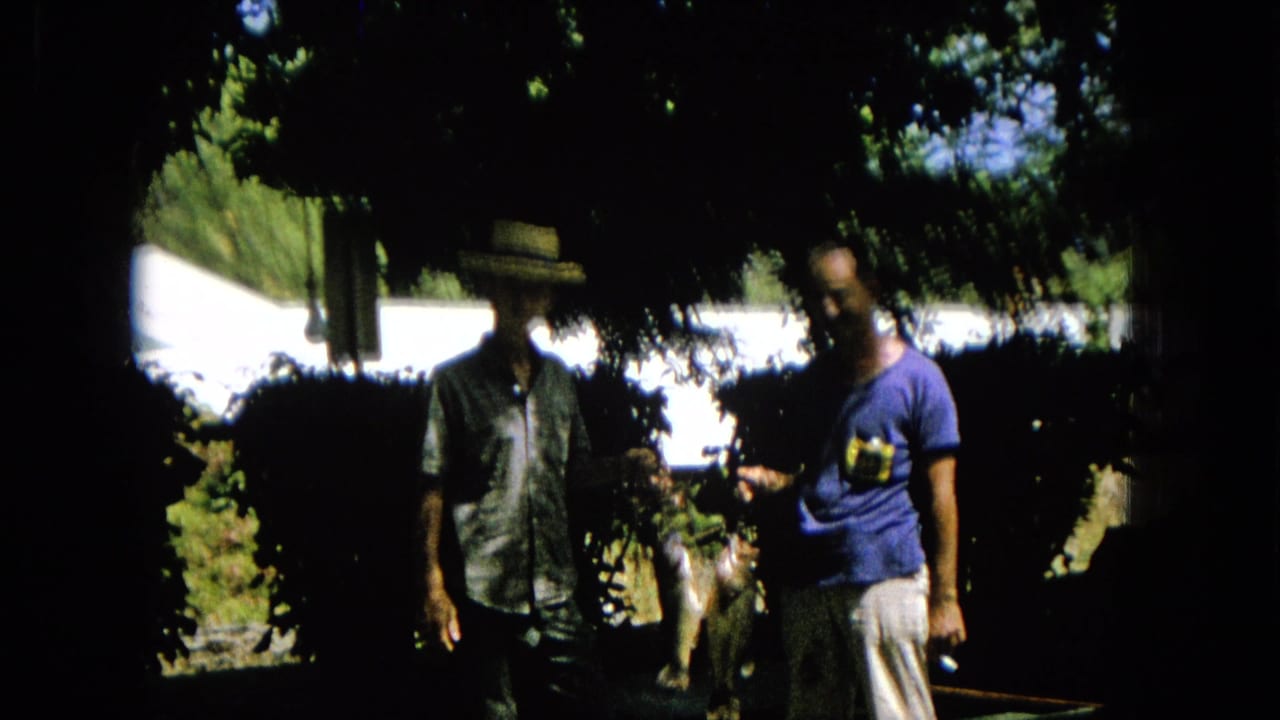 1965 8mm home-movie frame showing two adult men in a shaded outdoor yard or garden, with one in a straw hat and another in a blue t-shirt.
