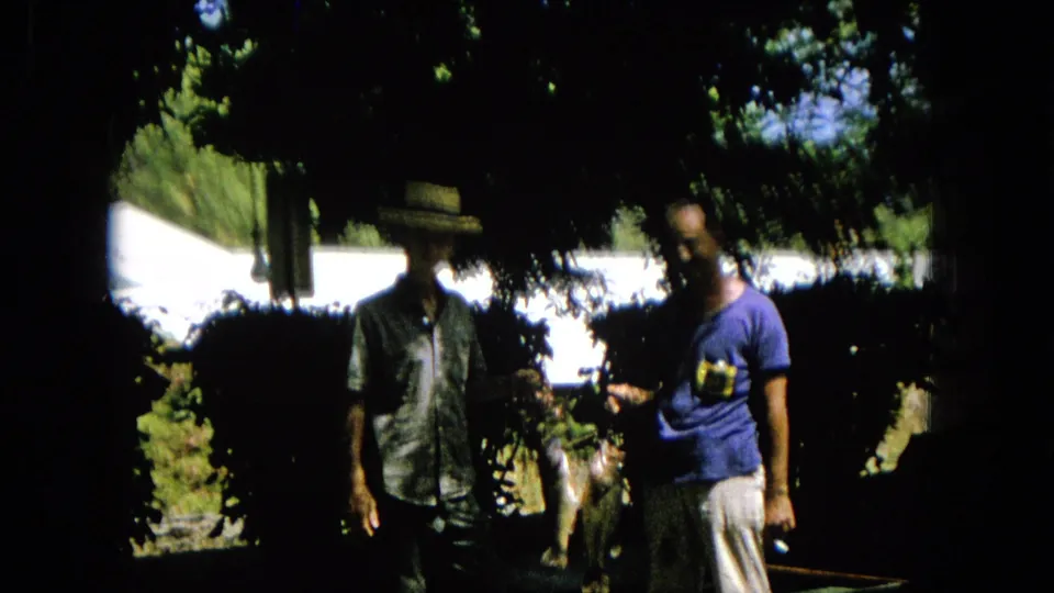 1965 8mm home-movie frame showing two adult men in a shaded outdoor yard or garden, with one in a straw hat and another in a blue t-shirt.